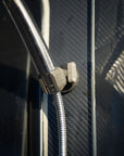 Tactic Engineering magnetic shower mount holding stainless steel shower head and flexible hose against carbon fiber van exterior, close up showing the SLS 3D printed nylon clip securely gripping the hose with neodymium magnet base on textured surface