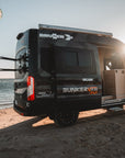 BunkerVan parked on beach with ocean and sky in the background, featuring Tactic Engineering magnetic kite carrier mounts on rear doors for kiteboarding adventures