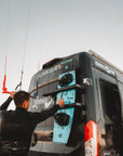 Person attaching a kiteboard to the rear doors of a BunkerVan using Tactic Engineering magnetic carrier mounts at a kitesurf beach location