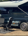 Fiat Ducato Carrera campervan with Thule awning extended using Tactic Engineering magnetic awning supports, campsite setup with folding chairs on dirt surface showing real world van life use