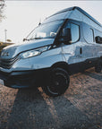Iveco Daily 4x4 campervan front three quarter view on gravel road with MaxTrax MK2 recovery boards mounted using Tactic Engineering magnetic quick release supports, silver off road expedition van with all terrain tires