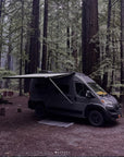 Ram ProMaster campervan parked in redwood forest with Thule awning extended using Tactic Engineering magnetic awning supports, showing campsite setup with outdoor mat and gear