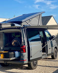 Volkswagen California T6 campervan rear view with Thule awning extended using Tactic Engineering magnetic awning supports, pop up roof raised and solar panels visible, open trunk with camping gear on gravel driveway