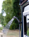 Tactic Engineering magnetic outdoor shower in use on white campervan, shower head magnetically mounted to van exterior with water flowing, green trees in background, vanlife outdoor shower setup from Belgium by @hobus_offgrid_buscampers