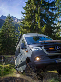 Mercedes Sprinter van with MaxTrax recovery boards magnetically mounted on curved bonnet using Tactic Engineering magnetic mounts for curved surfaces, off road adventure scene in mountain forest with pine trees and rocky peaks in background, headlights on at dusk