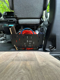 Tactic Engineering magnetic under seat organizer installed beneath Mercedes Sprinter 907 passenger seat, black MOLLE panel holding tools brushes and red extension cord, tool free magnetic mounting for 2019 to 2026 Sprinter models with wood floor visible
