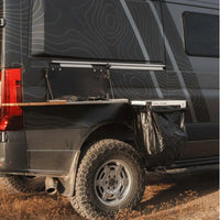 OWL Trail Trash magnetic camping trash holder mounted on rear of dark overland van holding a black trash bag, steel hoop base with neodymium magnets attached to vehicle body, off road tires and topographic wrap visible on expedition rig