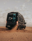 BunkerVan Ford Transit driving on desert dirt road with Duotone kiteboards magnetically secured to rear doors using Tactic Engineering magnetic carrier, kicking up sand and dust