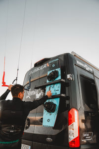 Person attaching a kiteboard to the rear doors of a BunkerVan using Tactic Engineering magnetic carrier mounts at a kitesurf beach location