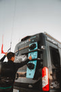 Person attaching a kiteboard to the rear doors of a BunkerVan using Tactic Engineering magnetic carrier mounts at a kitesurf beach location