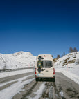 Camper van driving on snowy mountain road with Tactic Engineering magnetic ski carrier mounted on rear doors, carrying skis and snowboard in the Swiss Alps