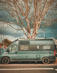 Fiat Ducato campervan with custom wrap parked under a tree, MaxTrax MK2 recovery boards mounted on roof using Tactic Engineering magnetic quick release supports, side view showing the adventure van setup
