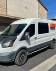 White Ford Transit van with orange MaxTrax MK2 recovery boards magnetically mounted on the side panel using Tactic Engineering magnetic quick release supports, parked in residential driveway showing the clean exterior mounting solution