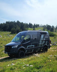Black Mercedes Sprinter campervan parked on grassy hillside with MaxTrax MK2 recovery boards mounted on roof rack using Tactic Engineering magnetic quick release supports, off road adventure setup with trees in background