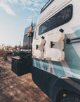 Mercedes Sprinter campervan with white RotoPax water containers magnetically mounted on side panel using Tactic Engineering magnetic RotoPax plate, desert landscape setting with outdoor cooking setup visible, overlanding adventure vehicle gear storage