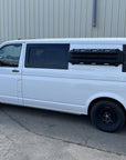 White Volkswagen Transporter T6 side view with black MaxTrax MK2 recovery boards mounted on rear window using Tactic Engineering magnetic quick release supports, rear spare tire carrier and roof rack visible